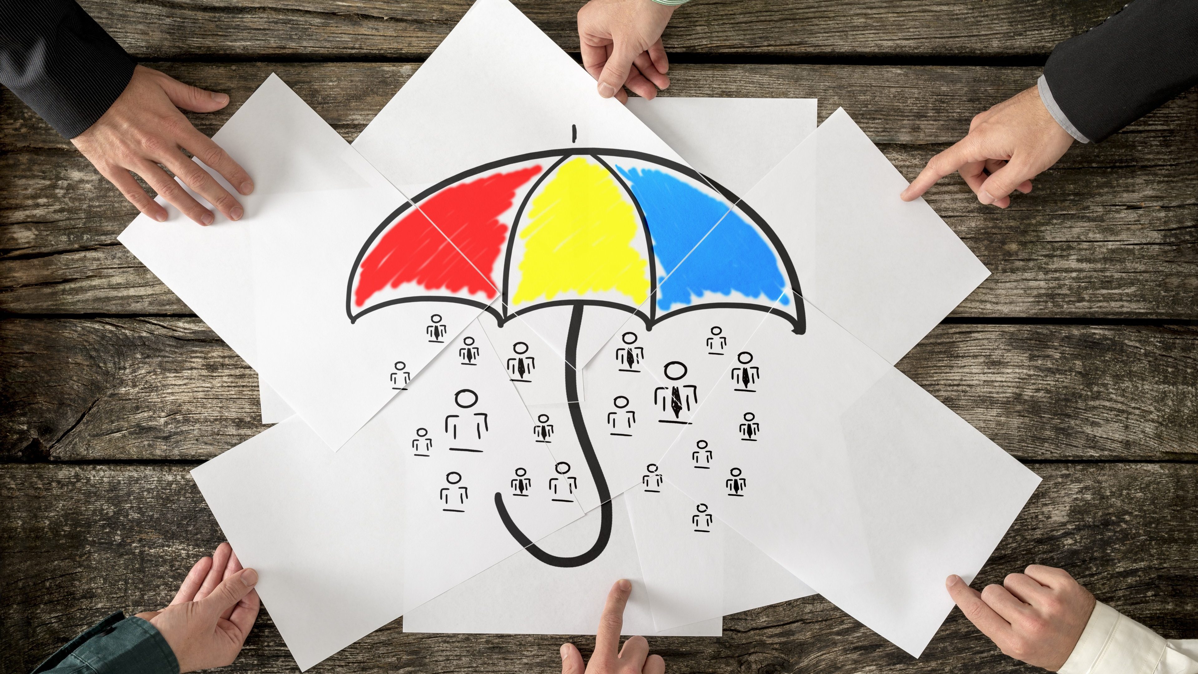 Safety and life insurance concept - six hands assembling a colourful umbrella sheltering many people icons drawn on white papers.